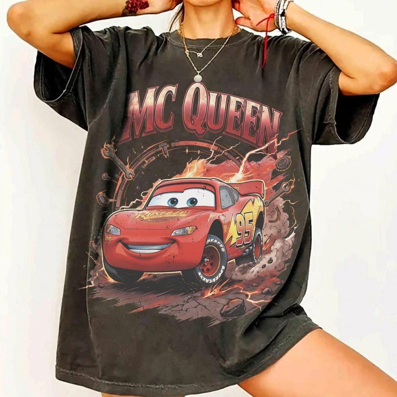 May include: Dark gray t-shirt featuring a graphic of Lightning McQueen from the movie Cars. The design includes the text "MC QUEEN" in red and a number "95" on the car. The shirt has a vintage, distressed look.