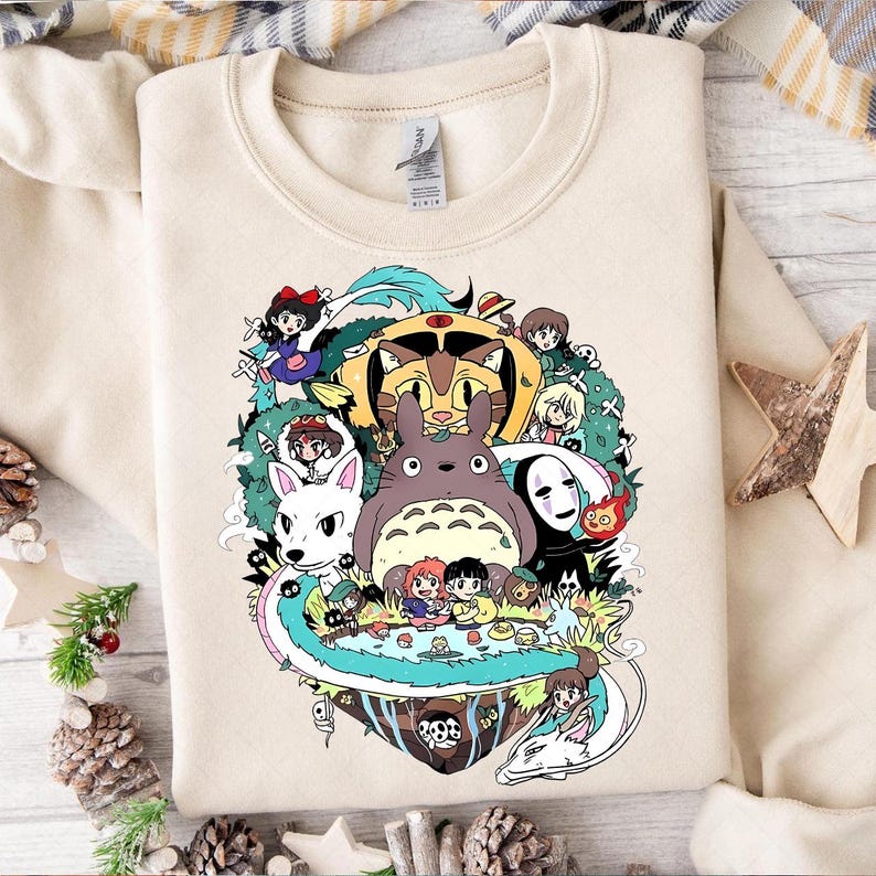 May include: A cream-colored sweatshirt with a colorful graphic print of characters from the Studio Ghibli film, 'My Neighbor Totoro'. The print features Totoro, Catbus, Kiki, and other characters from the film.