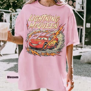 May include: A light pink t-shirt featuring a graphic of Lightning McQueen from the Cars movie. The design includes the text "LIGHTNING McQUEEN" above the car, with lightning bolts and a racing number 95.