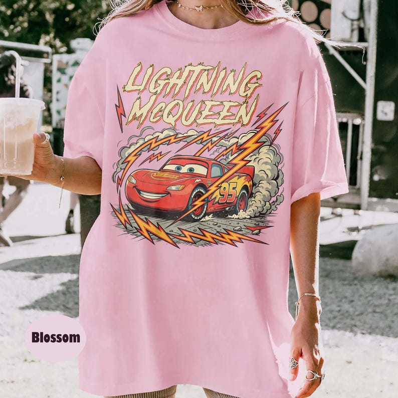 May include: A light pink t-shirt featuring a graphic of Lightning McQueen from the Cars movie. The design includes the text "LIGHTNING McQUEEN" above the car, with lightning bolts and a racing number 95.