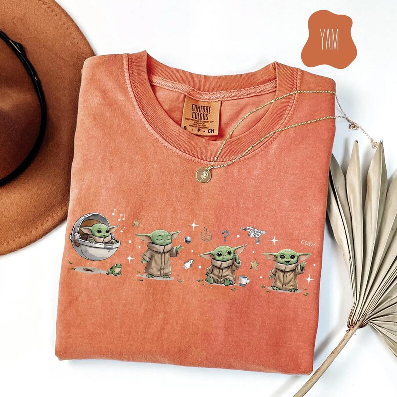 K&ouml;nnte beinhalten: Ein korallenfarbenes T-Shirt mit einer Grafik der Figur Grogu aus The Mandalorian. Das Design zeigt Grogu in verschiedenen Posen mit einem Frosch, einem Ball und einer Tasse. Das Shirt hat einen Rundhalsausschnitt und der Text "COOO" ist sichtbar.