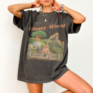 May include: Dark gray t-shirt featuring a colorful Disney World graphic. The graphic includes a castle, Spaceship Earth, and the text "Disney World" and "The most magical place on earth". The shirt is oversized with short sleeves.