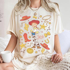 May include: Cream-colored t-shirt featuring a cartoon illustration of Jessie from Toy Story. The design includes a red hat, boots, sheriff badges, and the name "Jessie". Additional elements include cacti, stars, and a lasso.