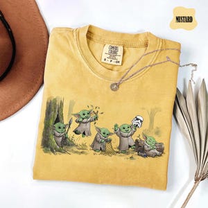 May include: A mustard yellow short-sleeve t-shirt with a crew neckline and a graphic print featuring five small green creatures in robes. One creature holds a spear, one uses a slingshot, one holds a net, one holds a helmet, and one sleeps.