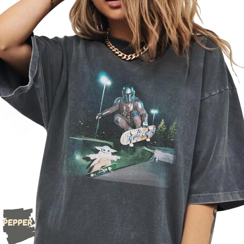 Op de afbeelding: Donkergrijs T-shirt met een afbeelding van The Mandalorian en Grogu die 's nachts skateboarden. Het ontwerp bevat een skateboardhelling, straatverlichting en bomen. Het shirt heeft een vintage, versleten uitstraling.