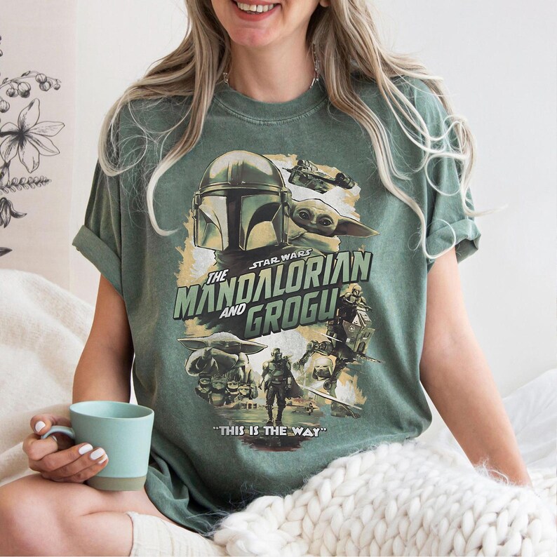 K&ouml;nnte beinhalten: Gr&uuml;nes T-Shirt mit einem Star Wars-Motiv mit "The Mandalorian und Grogu". Das Design enth&auml;lt Bilder von The Mandalorian, Grogu und anderen Charakteren mit dem Satz "THIS IS THE WAY" am unteren Rand.