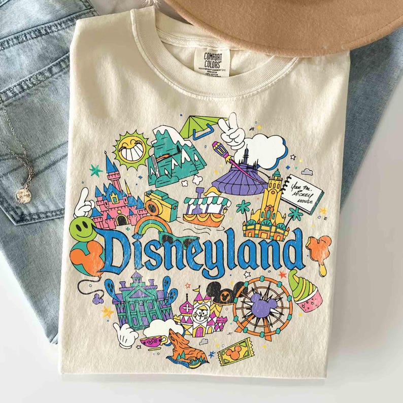 May include: Cream-colored crew neck sweatshirt with a colorful Disneyland graphic. The graphic features various park icons, including a castle, a Ferris wheel, and the Disneyland logo in blue. The sweatshirt is paired with blue jeans and a brown hat.