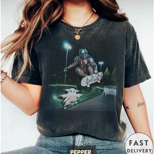 Op de afbeelding: Donkergrijze t-shirt met een afbeelding van The Mandalorian en Grogu die skateboarden in een skatepark. Het ontwerp toont de personages en skateboards tegen een nachtelijke sc&egrave;ne met lichten. Het t-shirt heeft een vintage uitstraling.