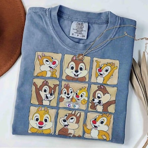 May include: A blue Comfort Colors t-shirt featuring a grid of Chip and Dale cartoon character illustrations. The shirt has a crew neck and short sleeves. A gold necklace with a pendant is draped across the shirt.