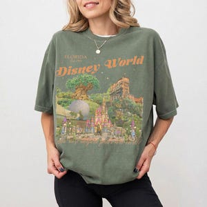 May include: Olive green t-shirt with a graphic of Disney World landmarks, including the Tree of Life, Cinderella Castle, and the Tower of Terror. The text "Disney World" and "Florida Est. 1971" are printed in orange. The phrase "the most magical place on earth" is also printed on the shirt.