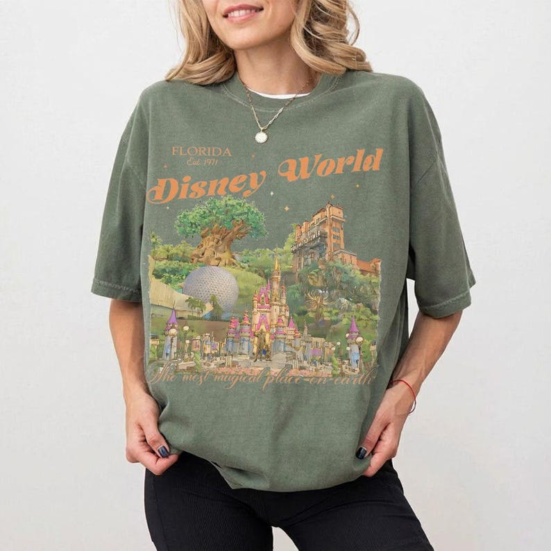 May include: Olive green t-shirt with a graphic of Disney World landmarks, including the Tree of Life, Cinderella Castle, and the Tower of Terror. The text "Disney World" and "Florida Est. 1971" are printed in orange. The phrase "the most magical place on earth" is also printed on the shirt.