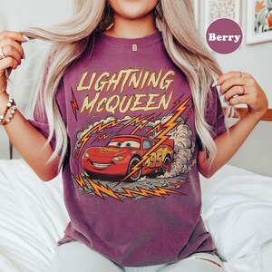 May include: Berry-colored t-shirt featuring a graphic of Lightning McQueen from the Cars movie. The design includes the text "Lightning McQueen" in yellow and red, along with a red race car with the number 95. The shirt has a vintage, distressed look.