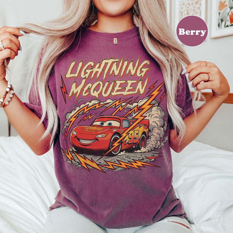 May include: Berry-colored t-shirt featuring a graphic of Lightning McQueen from the Cars movie. The design includes the text "Lightning McQueen" in yellow and red, along with a red race car with the number 95. The shirt has a vintage, distressed look.