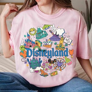 May include: Pink t-shirt with a colorful Disneyland graphic. The design includes a castle, a Ferris wheel, and the word "Disneyland" in blue. The shirt has short sleeves and a crew neck.