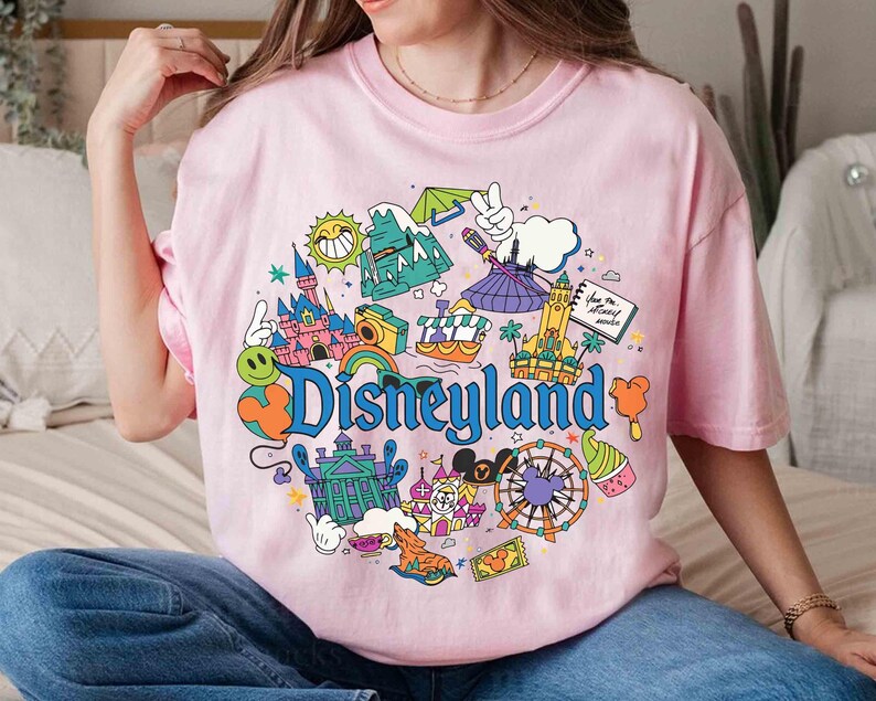 May include: Pink t-shirt with a colorful Disneyland graphic. The design includes a castle, a Ferris wheel, and the word "Disneyland" in blue. The shirt has short sleeves and a crew neck.