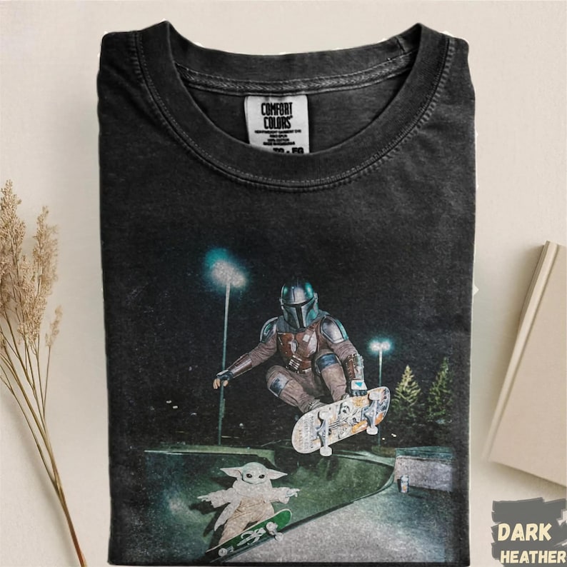 Op de afbeelding: Donker heather t-shirt met een afbeelding van The Mandalorian aan het skateboarden, met Grogu ook op een skateboard. Het ontwerp speelt zich af in een skatepark 's nachts. Het shirt heeft een Comfort Colors label.