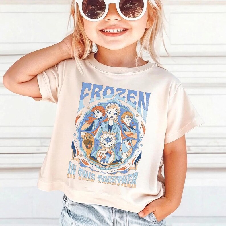 May include: A light beige t-shirt featuring the word "FROZEN" in blue, with a graphic of characters from the movie. The text "IN THIS TOGETHER" is below the graphic. The shirt is short-sleeved.