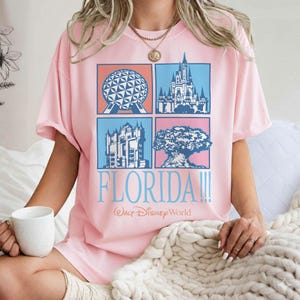 May include: Pink t-shirt featuring four square images of Florida landmarks. The shirt has the word "FLORIDA!!!" in light blue, with "Walt Disney World" below. The shirt is short-sleeved and has a relaxed fit.