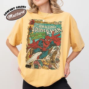 Puede incluir: Una camiseta de color amarillo mostaza con un gr&aacute;fico de portada de c&oacute;mic vintage de Marvel, The Amazing Spider-Man. El estampado muestra a Spider-Man con su traje rojo y azul, junto al texto "The Amazing Spider-Man" y "Comfort Colors Mustard".