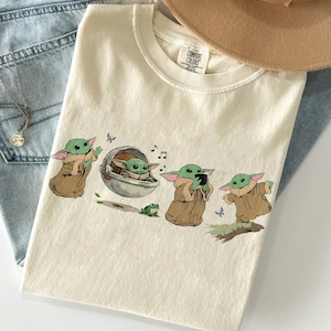 May include: Cream-coloured t-shirt with a cartoon illustration of a Grogu character in different poses. The design includes musical notes, butterflies, and a small green frog. The shirt is folded on top of blue jeans.