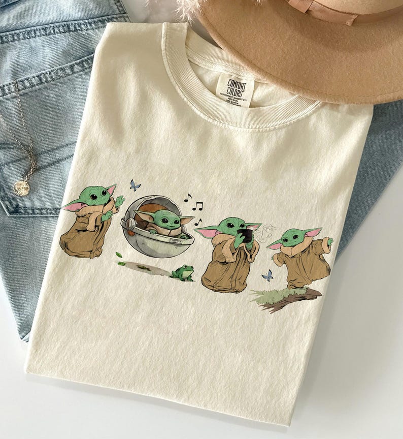 May include: Cream-coloured t-shirt with a cartoon illustration of a Grogu character in different poses. The design includes musical notes, butterflies, and a small green frog. The shirt is folded on top of blue jeans.