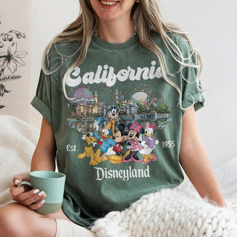 Może przedstawiać: Zielona koszulka z grafiką Disneylandu. Grafika przedstawia słowo "California" nad kolorową ilustracją zabytk&oacute;w i postaci z Disneylandu, w tym Myszki Miki i Minnie. Tekst "Est. 1955" i "Disneyland" są r&oacute;wnież uwzględnione.
