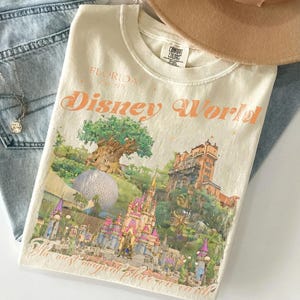 May include: Cream-colored t-shirt with a Disney World graphic. The graphic features a castle, tree, and other park landmarks. The text "Florida Est. 1971" and "Disney World" are printed in orange. A tan hat and blue jeans are in the background.