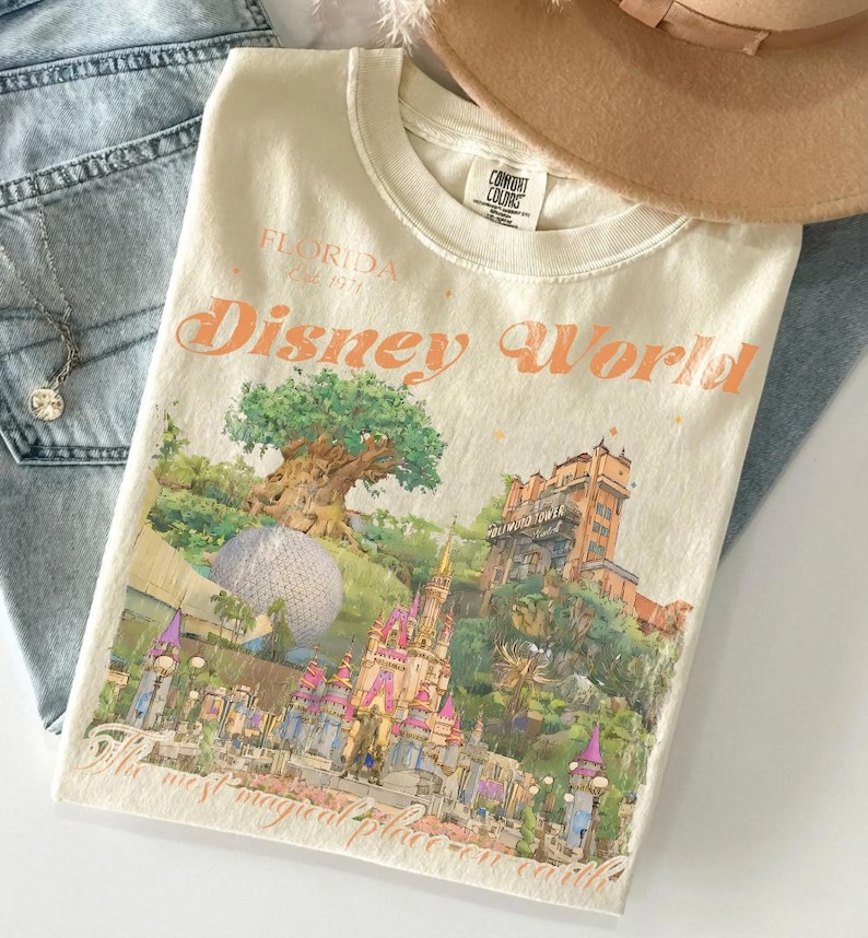 May include: Cream-colored t-shirt with a Disney World graphic. The graphic features a castle, tree, and other park landmarks. The text "Florida Est. 1971" and "Disney World" are printed in orange. A tan hat and blue jeans are in the background.