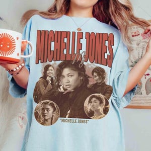 May include: Light blue t-shirt featuring the name "MICHELLE JONES" in a retro font. The shirt includes multiple images of a woman, with the text "MICHELLE JONES" below the images. A white mug with a sun design is held in the image.