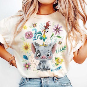 K&ouml;nnte beinhalten: Cremefarbenes T-Shirt mit einem Cartoon-Schwein mit einem Hahn auf dem Kopf, umgeben von bunten Blumen und Bl&auml;ttern. Das Schwein ist grau mit einer rosa Schnauze und Ohren. Der Hahn ist gelb, blau und rot. Blumendesign.