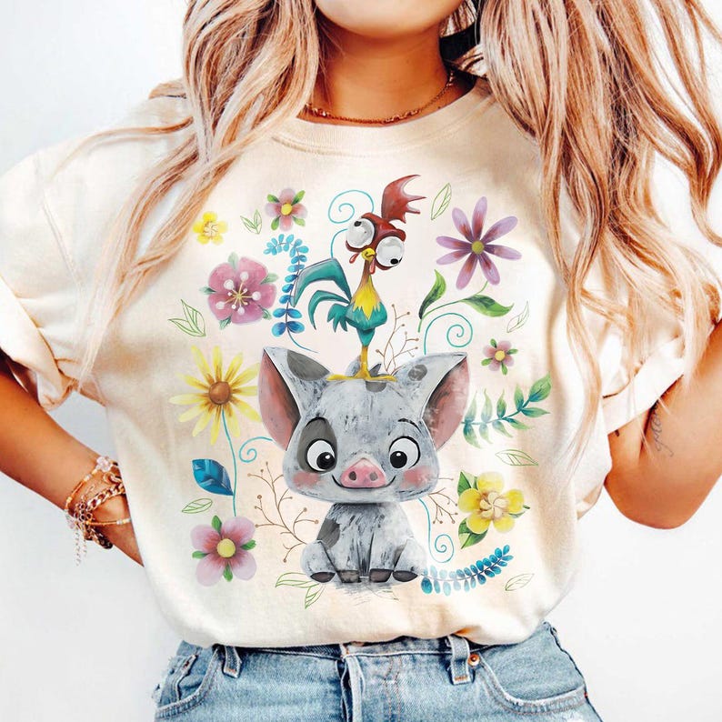 K&ouml;nnte beinhalten: Cremefarbenes T-Shirt mit einem Cartoon-Schwein mit einem Hahn auf dem Kopf, umgeben von bunten Blumen und Bl&auml;ttern. Das Schwein ist grau mit einer rosa Schnauze und Ohren. Der Hahn ist gelb, blau und rot. Blumendesign.
