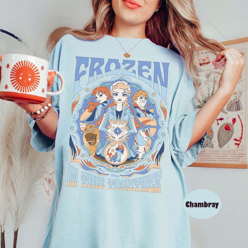 May include: Light blue t-shirt featuring the word "FROZEN" in a retro font, with a circular graphic of characters from the movie. The phrase "IN THIS TOGETHER" is printed below. A white mug with a sun design is held in the hand.