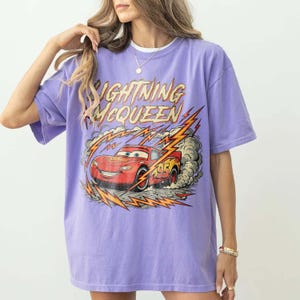 May include: A lilac-colored t-shirt featuring a graphic of Lightning McQueen from the Cars movie. The design includes the text "Lightning McQueen" in yellow and red, with the number 95 on the car. The car is depicted with lightning bolts and smoke.