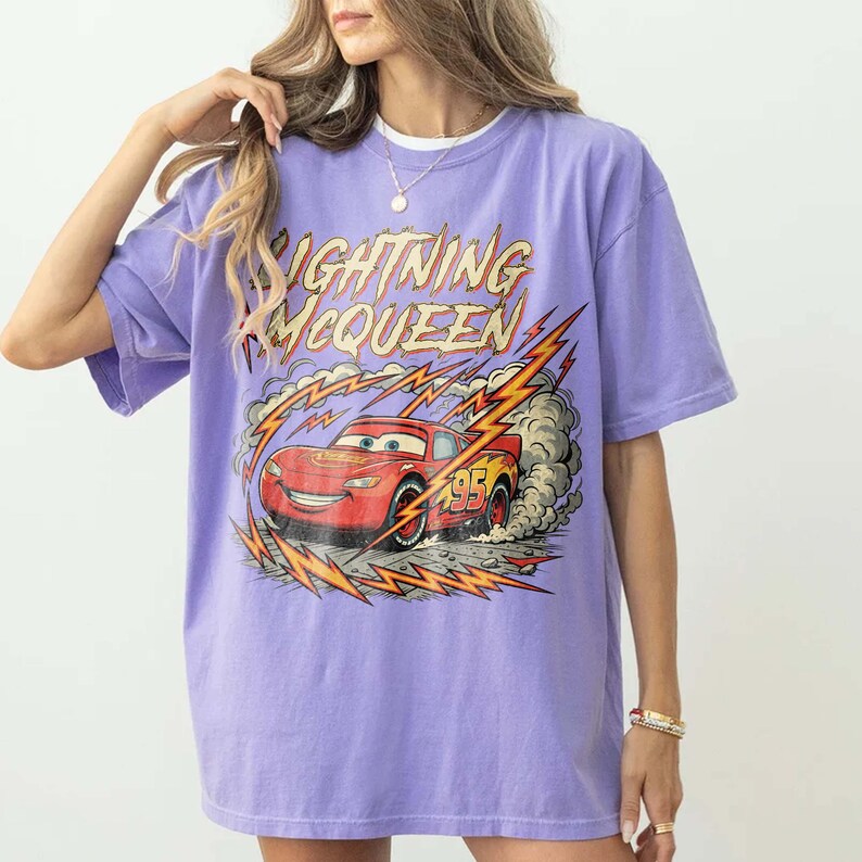May include: A lilac-colored t-shirt featuring a graphic of Lightning McQueen from the Cars movie. The design includes the text "Lightning McQueen" in yellow and red, with the number 95 on the car. The car is depicted with lightning bolts and smoke.
