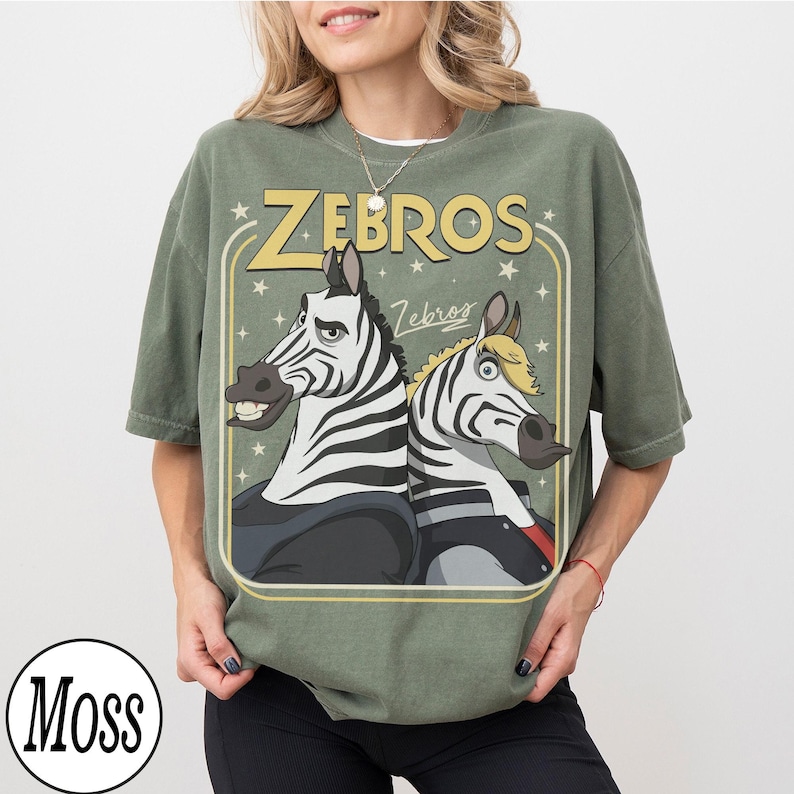 May include: A moss green t-shirt featuring a cartoon graphic of two zebras with the text "ZEBROS" in yellow. The zebras are wearing jackets and have expressive faces. The shirt has a vintage, slightly faded look.