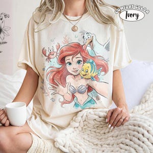 Op de afbeelding: Een ivoorkleurig T-shirt met korte mouwen en een grafische print van een zeemeermin met rood haar, een gele en blauwe vis, een rode krab en een witte meeuw. De tekst "COMFORT COLOR IVORY" staat rechtsboven in het ontwerp gedrukt.