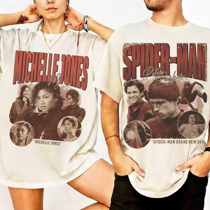May include: Two cream-colored t-shirts featuring movie graphics. One shirt displays "MICHELLE JONES" with images of the actress. The other shirt says "SPIDER-MAN Peter Parker" with images of the actor. Both shirts have a vintage style.