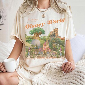 May include: Cream-colored t-shirt featuring a colorful Disney World graphic. The design includes the text "Florida Est. 1971" and "Disney World" in orange, along with illustrations of the park's landmarks. The shirt also has the text "The most magical place on earth."