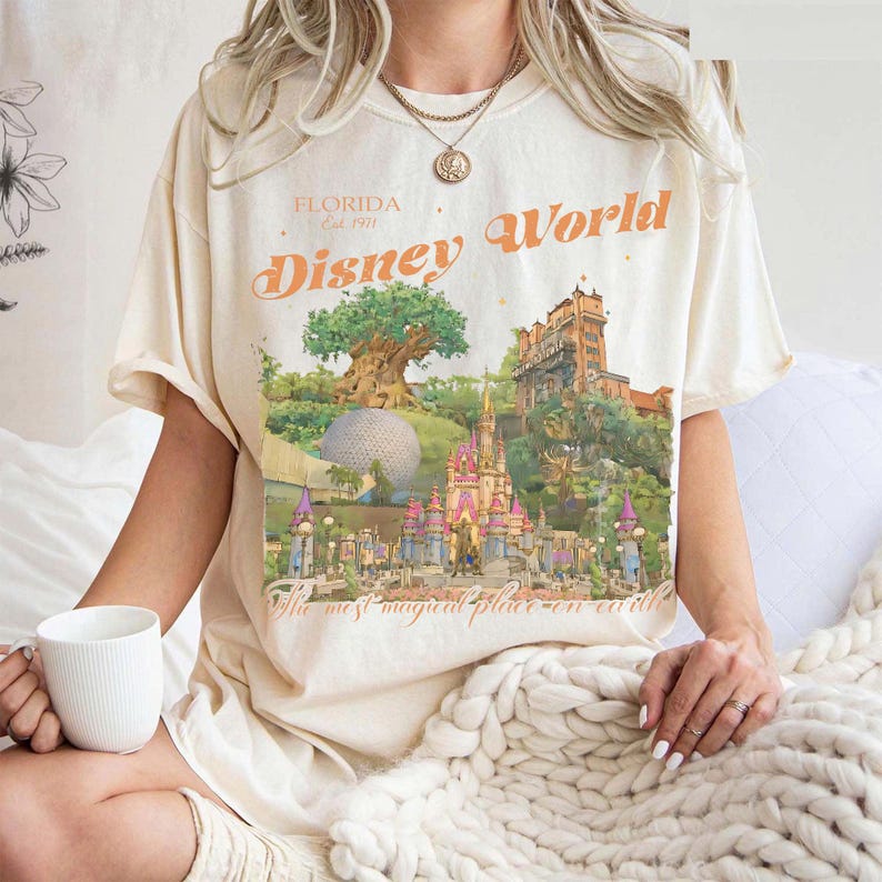 May include: Cream-colored t-shirt featuring a colorful Disney World graphic. The design includes the text "Florida Est. 1971" and "Disney World" in orange, along with illustrations of the park's landmarks. The shirt also has the text "The most magical place on earth."
