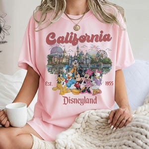 Może przedstawiać: R&oacute;żowa koszulka z nadrukiem Disneyland. Projekt zawiera napis "California" w stylu retro, zamek Disneyland i postacie z kresk&oacute;wek. Tekst "Est. 1955" i "Disneyland" są r&oacute;wnież uwzględnione.