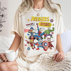 May include: Cream-colored t-shirt featuring a graphic of Disney characters as Avengers superheroes. The design includes Mickey Mouse, Minnie Mouse, Donald Duck, and others in superhero poses, with the text "The Avengers" and "Avengers Assemble!"