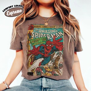 Puede incluir: Una camiseta de manga corta de color marr&oacute;n oscuro con un gr&aacute;fico de estilo vintage de la portada de un c&oacute;mic de Marvel, The Amazing Spider-Man. El estampado muestra a Spider-Man en una pose din&aacute;mica con colores rojo y azul.