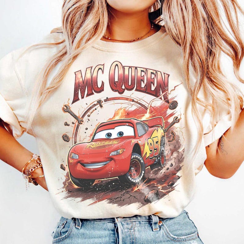 May include: Cream-colored t-shirt featuring a graphic of the animated character Lightning McQueen from the movie Cars. The design includes the text "MC QUEEN" above the car and the number "95" on the side. The car is red with yellow accents.