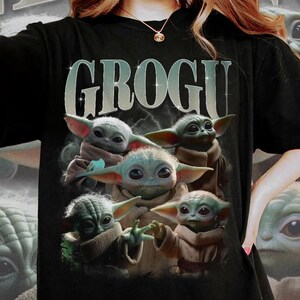 Puede incluir: Camiseta negra con la palabra "GROGU" en una fuente degradada sobre un gr&aacute;fico de cinco personajes Grogu. Los personajes Grogu tienen piel verde, orejas grandes y t&uacute;nicas marrones. El dise&ntilde;o tiene un estilo de arte digital.