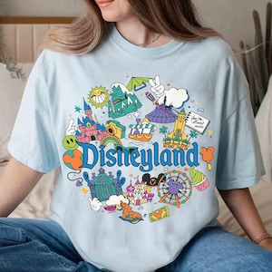 May include: Light blue t-shirt featuring a colorful Disneyland graphic. The design includes a rainbow, castle, and the word "Disneyland" in a playful font. The shirt is a comfortable, casual style.