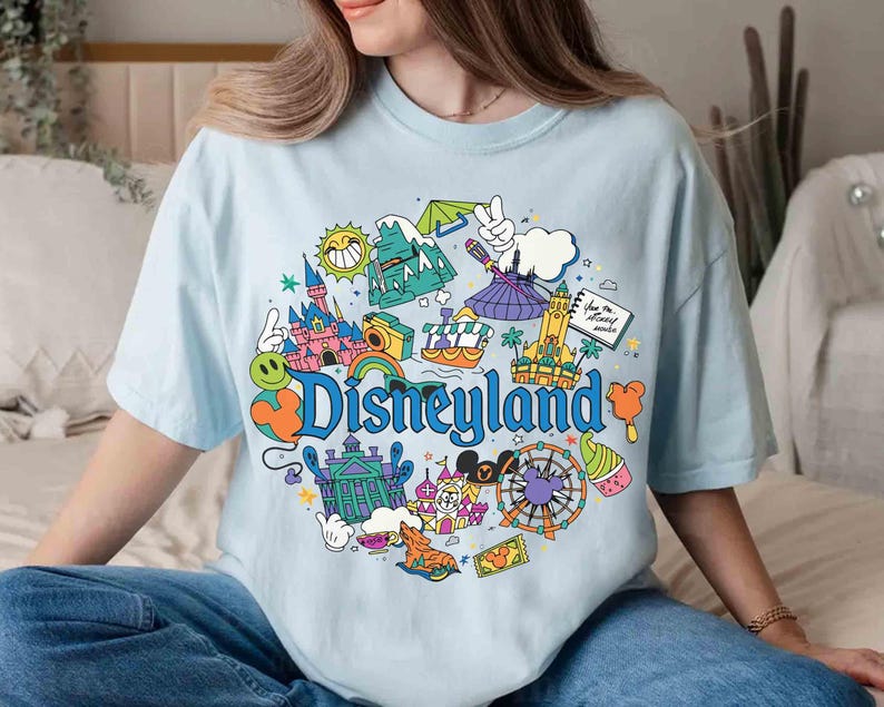 May include: Light blue t-shirt featuring a colorful Disneyland graphic. The design includes a rainbow, castle, and the word "Disneyland" in a playful font. The shirt is a comfortable, casual style.