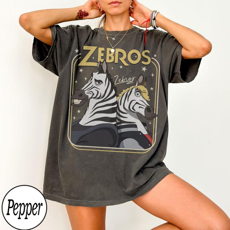 May include: Dark gray t-shirt featuring a cartoon illustration of two zebras with the text "ZEBROS" in a gold-colored frame. The zebras have black and white stripes and are set against a dark background with stars.
