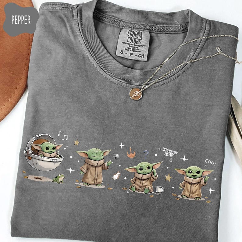 K&ouml;nnte beinhalten: Ein graues Comfort Colors T-Shirt mit einer Grogu-Grafik in verschiedenen Posen. Das Shirt hat einen Rundhalsausschnitt und ein kleines Etikett. Eine silberne Halskette mit Anh&auml;nger ist ebenfalls sichtbar.