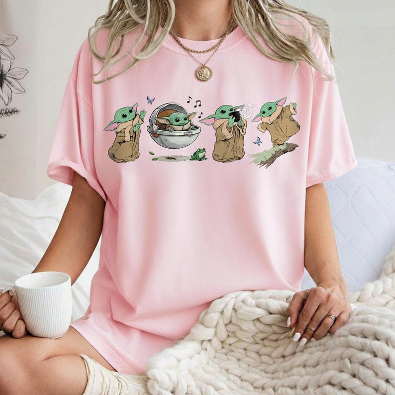 May include: Pale pink t-shirt with a cartoon graphic of a character from a popular science fiction series. The design shows the character in different poses, with musical notes and a frog. The shirt is a light pink colour.