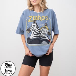 May include: A blue t-shirt featuring a cartoon graphic of two zebras with the word "ZEBROS" in yellow. The zebras are in a rectangular frame with stars. The shirt is a light blue color.